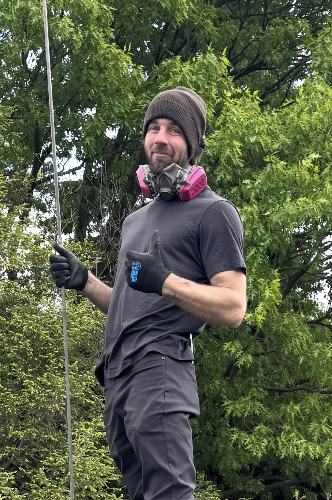 Sam on a chimney in a beanie and work gloves, respirator around his neck, holding a sweep rod and giving a thumbs-up.
